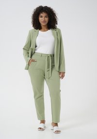 Un costume vert clair avec une veste cintrée et un pantalon fuselé, doté d'une ceinture à nouer. Porté sur un haut blanc et associé à des sandales à talons blanches.