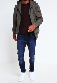 Huva grön jacka, vinröd henley-tröja, blå rivna jeans och vita sneakers. Avslappnad passform med texturerad detalj på tröjan.