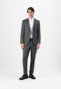 Férfi áll egy fehér falnak támaszkodva, szürke öltönyben, fehér gombos ingben és fekete dresszcipőben, előre nézve, lazán álló testtartásban.
