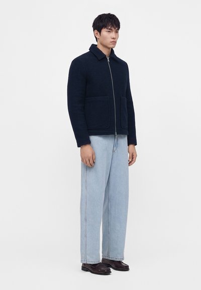 Homme portant une veste zippée bleu marine avec des poches devant, un jean large bleu clair et des chaussures en cuir marron foncé, debout devant un fond uni.