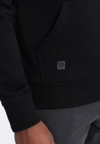 Sudadera negra con dobladillo y puños acanalados, con un bolsillo frontal y un pequeño parche de logo cuadrado cerca del bolsillo.