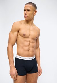 Μυώδης άνδρας που φοράει μαύρα μποξέρ Tommy Hilfiger με λευκή μέση, στέκεται μπροστά σε απλό ανοιχτό φόντο, κοιτάζοντας αριστερά.