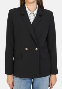 Blazer negro de doble botonadura con solapas con muesca, botones en tono dorado y dos bolsillos frontales, combinado con una camisa blanca a cuadros.