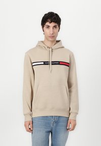Bežinė džinsinė su priekinėmis kišenėmis, su raišteliu, ir grafika, kurioje yra juodos, baltos ir raudonos juostelės su užrašu "TOMMY HILFIGER".