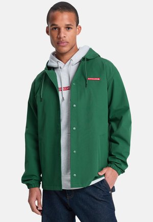 Jonge man die een groene capuchonjas draagt over een lichtgrijze Quiksilver-hoodie en donkerblauwe jeans, staand met één hand in de zak.