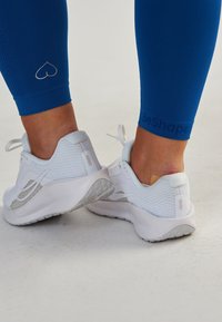Leggings de sport bleus avec un logo en forme de cœur et la marque "BeShaped", associés à des baskets blanches texturées avec des accents gris et des lacets.