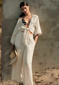 Lichtbeige linnenoutfit met een geknoopt blouse en wijdvallende broek. Een zwart-wit gestreepte bikini eronder en een rieten hoed in de hand.