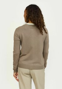 Smeđi pleteni pulover s okruglim izrezom i rebrastim manžetama, nosi se preko bež hlača. Ima glatku teksturu i uski kroj.