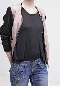 Lichtroze bomberjack met ritssluiting en zwarte stoffen mouwen, gedragen over een zwart T-shirt en donkerblauwe jeans met een uniek zakontwerp.