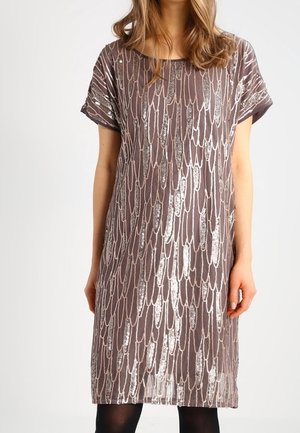 Vrouw die een mauve jurk tot op de knie draagt met een zilveren paillettenkleurig verenpatroon en korte opgerolde mouwen, staand tegen een witte achtergrond.