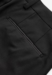 Záber zblízka na čiernu šitú časť látky s horizontálnym klopovým vreckom s precíznym prešitím a pútkom na opasok nad.