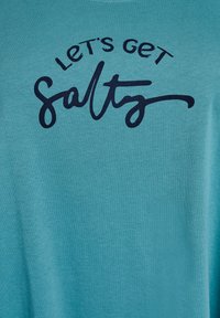 Turkusowa koszulka z bawełny z czarnym kursywą tekstem: "Let's get Salty" w półokrągłym układzie wokół słowa "Salty."