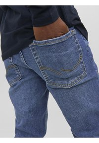 Bleka blå denimjeans med gula sömmar, med två bakfickor med böjda mönster, visade från sidan med en hand i fickan.