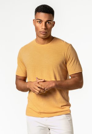 Junger Mann mit kurzen Haaren, der ein gelbes kurzärmeliges Hemd und weiße Hosen trägt und mit verschränkten Händen vor einem einfachen Hintergrund steht.