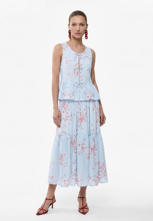 Femme portant une robe sans manches bleu clair à fleurs avec jupe à volants, détails noués à l'avant, boucles d'oreilles rouges et talons rouges à lanières, debout devant un fond blanc.