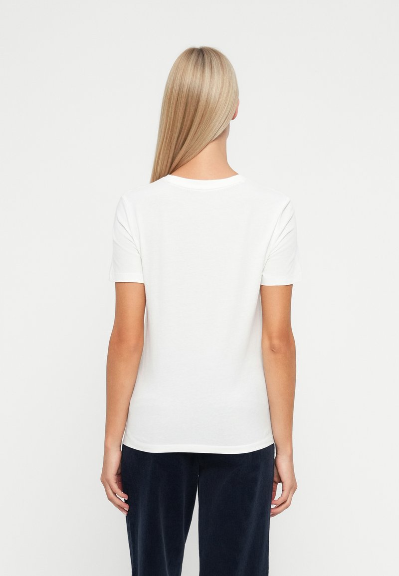 Blonde Frau von hinten gesehen, die ein schlichtes weißes T-Shirt mit kurzen Ärmeln und dunkelblauen Hosen vor einem weißen Hintergrund trägt.