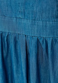 Denim jurk met een rijke blauwe kleur, geweven stof en geplooide details bij de taille, wat een vloeiende rok effect creëert.