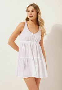 Witte mouwloze jurk met een ronde hals, geplooide taille en gelaagde rok. Heeft decoratieve bruine stiksels langs de halslijn.