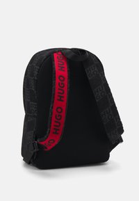 Zaino nero con finitura testurizzata, caratterizzato da una strap rossa prominente con il marchio "HUGO". Pannelli laterali in rete per la ventilazione. Spallacci regolabili.