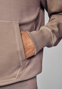 Mikina v tlmenej taupe farbe, vyrobená z mäkkej, textúrovanej látky, s rebrovanými manžetami a detailom klokanieho vrecka.