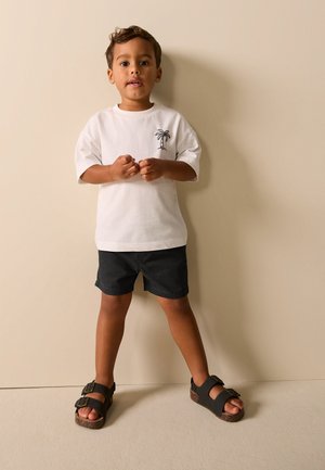 Jeune garçon debout contre un mur beige, portant un t-shirt blanc avec un motif de palmier, un short noir et des sandales noires à boucles.