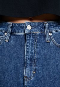 Närbild av midjan med blå denimjeans som är knäppta, som visar bältesöglor, sömnadsdetaljer och en mörk övre fåll ovanför en liten synlig navel.