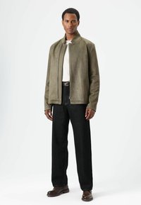 Olivgrüne Wildlederjacke mit Stehkragen und Reißverschluss vorne, kombiniert mit schwarzen Jeans mit weitem Bein und braunen Lederschuhen.