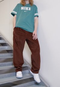 Teal Nike-t-shirt med vitt märke och ränder, i kombination med bruna manchesterbyxor. Modellen står i vita sneakers på metalltrappor.