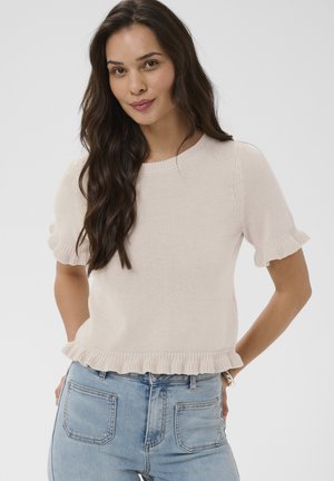 Donna con capelli lunghi e scuri che indossa un top lavorato a maglia beige a maniche corte con bordi arricciati e jeans azzurri chiari, in piedi davanti a uno sfondo semplice.