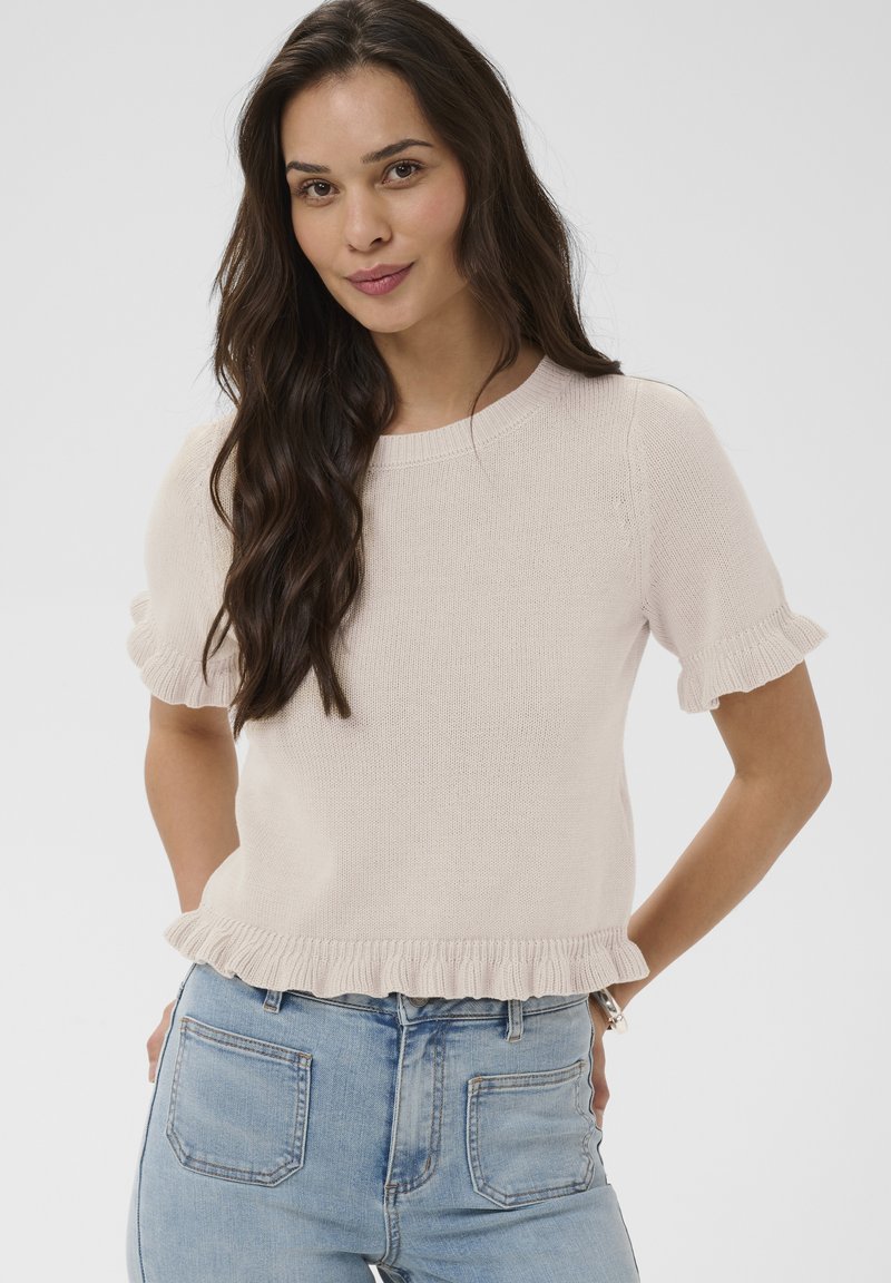 Femme aux longs cheveux foncés portant un haut en tricot beige à manches courtes avec des bords volantés et un jean bleu clair, debout devant un fond uni.