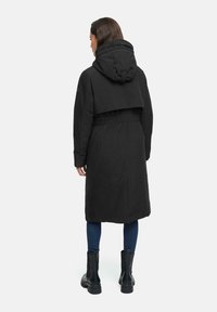 Black long coat with a cinched waist and a large hood. Features an embroidered logo on the back and is made of textured fabric.