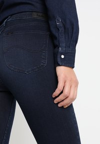 Jeans de mezclilla azul oscuro con un corte ajustado. Las características incluyen un diseño distintivo en el bolsillo trasero, costuras sutiles y un logo en la cinturilla.