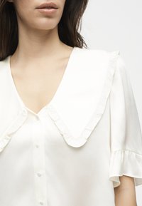 Γυναίκα που φοράει λευκό μπλουζάκι με μεγάλο μυτερό γιακά και βολάν περιμετρικά, κλειστό με κουμπιά μπροστά και κοντά μανίκια με βολάν στις μανσέτες.