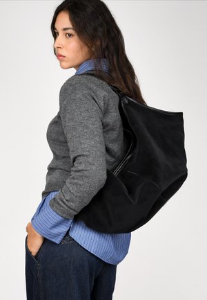 Mochila de cuero negra con textura suave, forma relajada y diseño minimalista. Lleva sobre el hombro con un suéter gris y una camisa de rayas azules.