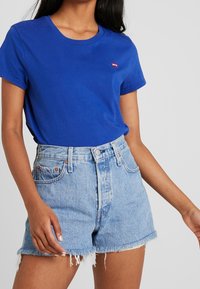 Blå bomullströja med korta ärmar, med en röd logotyp på bröstet, i kombination med högmidjade urblekta jeansshorts med fransad kant.