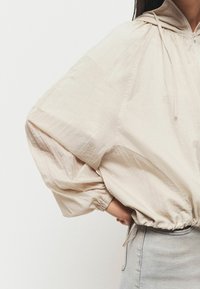Pull à capuche beige clair avec une finition texturée, des manches oversize et une taille à cordon élastique. Porté avec une jupe en denim clair.