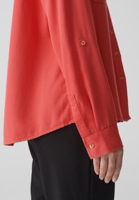 Camicia a maniche lunghe corallo con polsini abbottonati, dotata di orlo dritto e un taschino sul petto. Il tessuto ha una consistenza liscia.