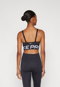 Fekete sportmelltartó állítható pántokkal, széles gumival, amelyen vastag fehér betűkkel olvasható a "NIKE PRO" felirat; magas derekú fekete leggings-szel párosítva.