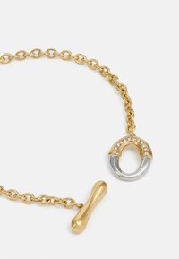 Goldene Kettenarmband mit einem runden Anhänger, der silberne Akzente und Strassdetails aufweist, sowie einem kleinen, verlängerten goldenen Verschluss.