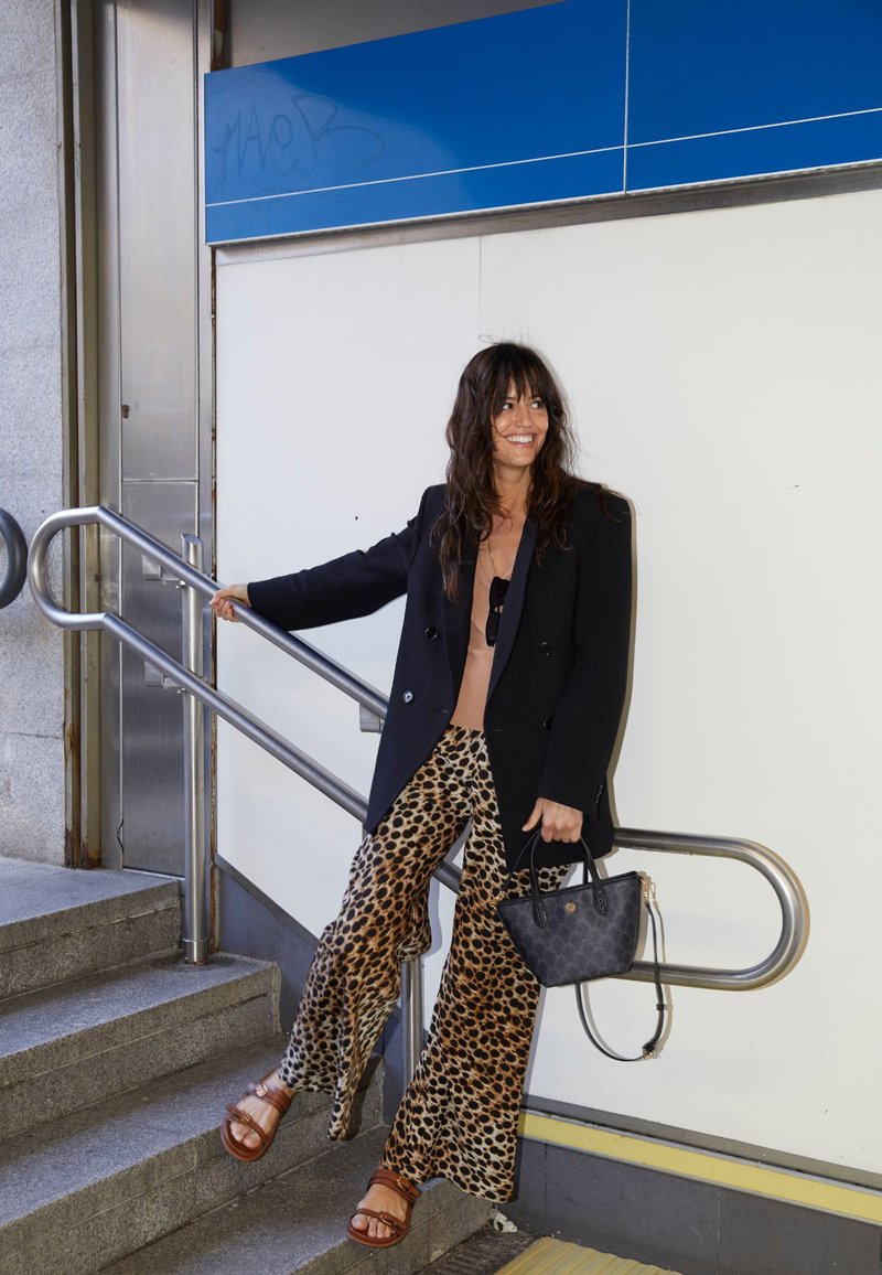 Czarna marynarka dwurzędowa na beżowej bluzce, zestawiona z spodniami w panterkę o szerokich nogawkach. Brązowe sandały i mała czarna torebka. Widoczny metalowy poręcz.