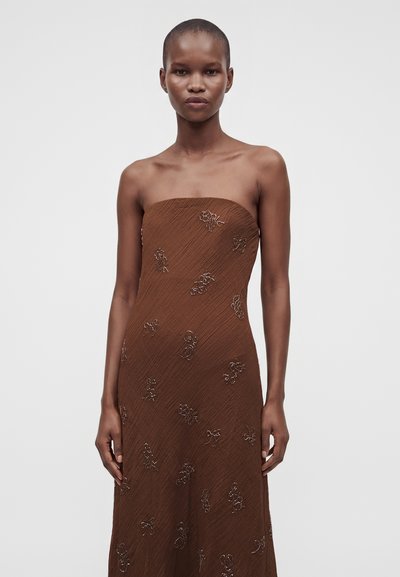 Femme portant une robe marron bustier avec une broderie florale subtile, debout devant un fond clair uni.