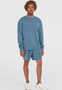 Blå sweatshirt med rund halsudskæring og logoemblem, kombineret med matchende shorts med mønstrede sidepaneler og hvide sneakers.