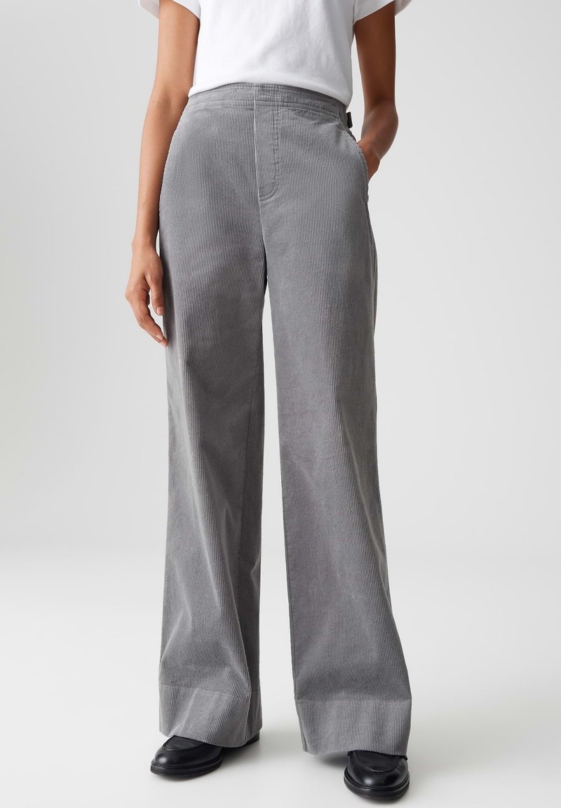 Personne portant un pantalon large en velours côtelé gris avec des poches latérales et des chaussures noires, debout devant un fond uni.