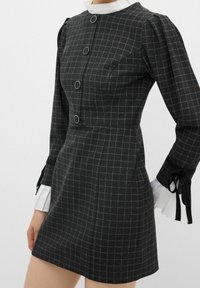 Robe à carreaux gris foncé avec manches longues bouffantes, grands boutons ronds sur le devant, poignets blancs à volants et liens en ruban noir aux poignets.