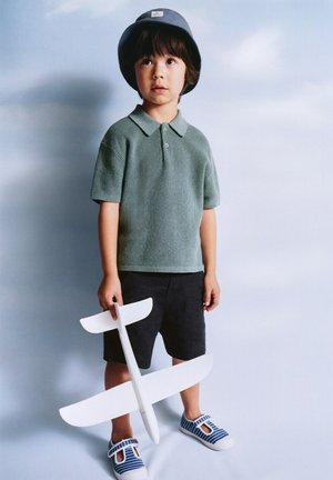 Jonge jongen met een emmerhoed, groen gebreid poloshirt, zwarte korte broek en gestreepte schoenen, die een wit speelgoedzweefvliegtuig vasthoudt tegen een lichte achtergrond.