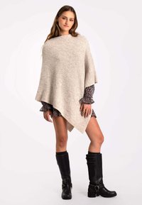 Poncho de punto beige con un dobladillo asimétrico y textura suave, superpuesto a un vestido estampado; acompañado de botas negras hasta la rodilla.