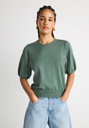 Νεαρή γυναίκα με πλεξούδες στα μαλλιά, φορώντας χρυσούς κρίκους στα αυτιά, ένα πράσινο πλεκτό μπλουζάκι με κοντά μανίκια και ανοιχτό μπλε τζιν με τα χέρια στις τσέπες.