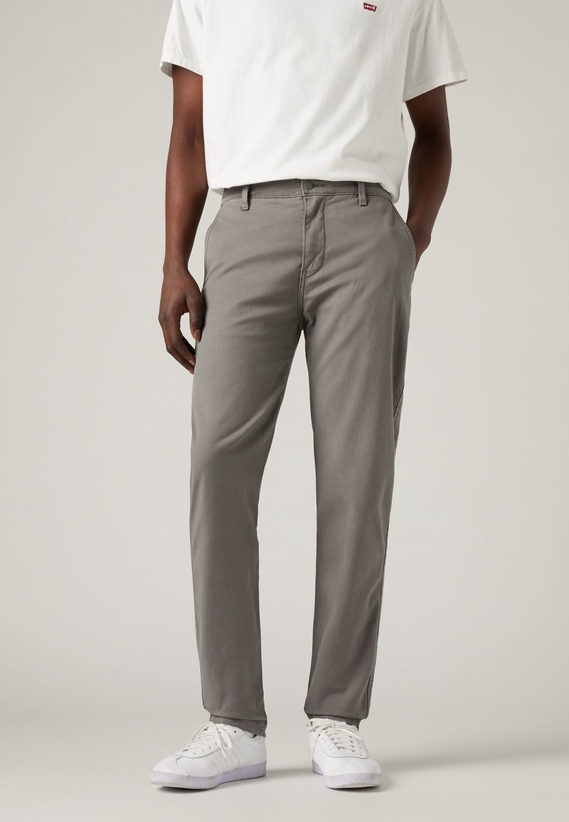 Homme portant un pantalon gris, des baskets blanches et un T-shirt blanc avec un petit logo rouge sur la poitrine, debout devant un fond uni.