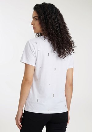 Donna con capelli ricci e scuri che indossa una maglietta bianca a maniche corte con piccole borchie argentate sparse, vista di spalle.