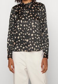 Blusa nera in satin con un motivo a pois beige chiaro, maniche lunghe con polsini abbottonati e chiusura frontale con bottoni.